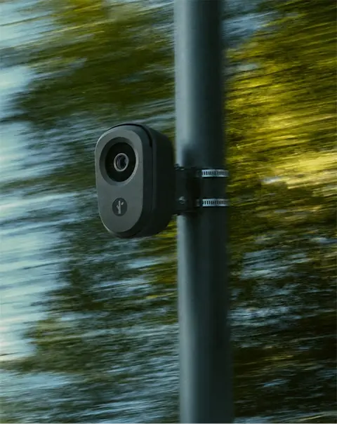 Black surveillance camera mounted on a pole with blurred green trees in the background.