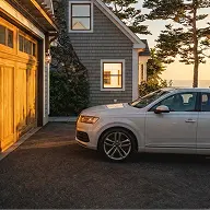 White SUV parked beside a house with gray siding and wooden garage door during sunset.