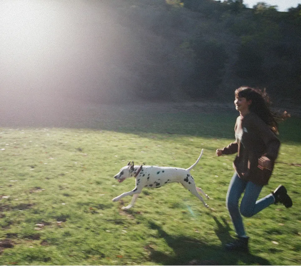 Woman running joyfully on grass alongside a running Dalmatian dog in sunlight.