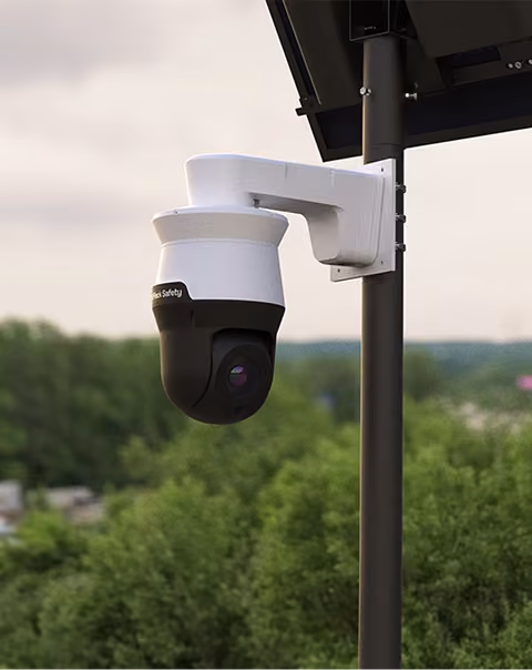 White and black security camera mounted on a pole with greenery and cloudy sky background.