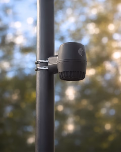 Black outdoor speaker mounted on a pole with metal straps against a blurred natural background.