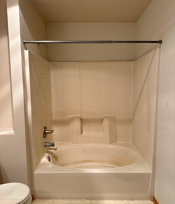 Outdated bathroom with an old built-in bathtub, white tile surround, and worn flooring before a tub to shower conversion