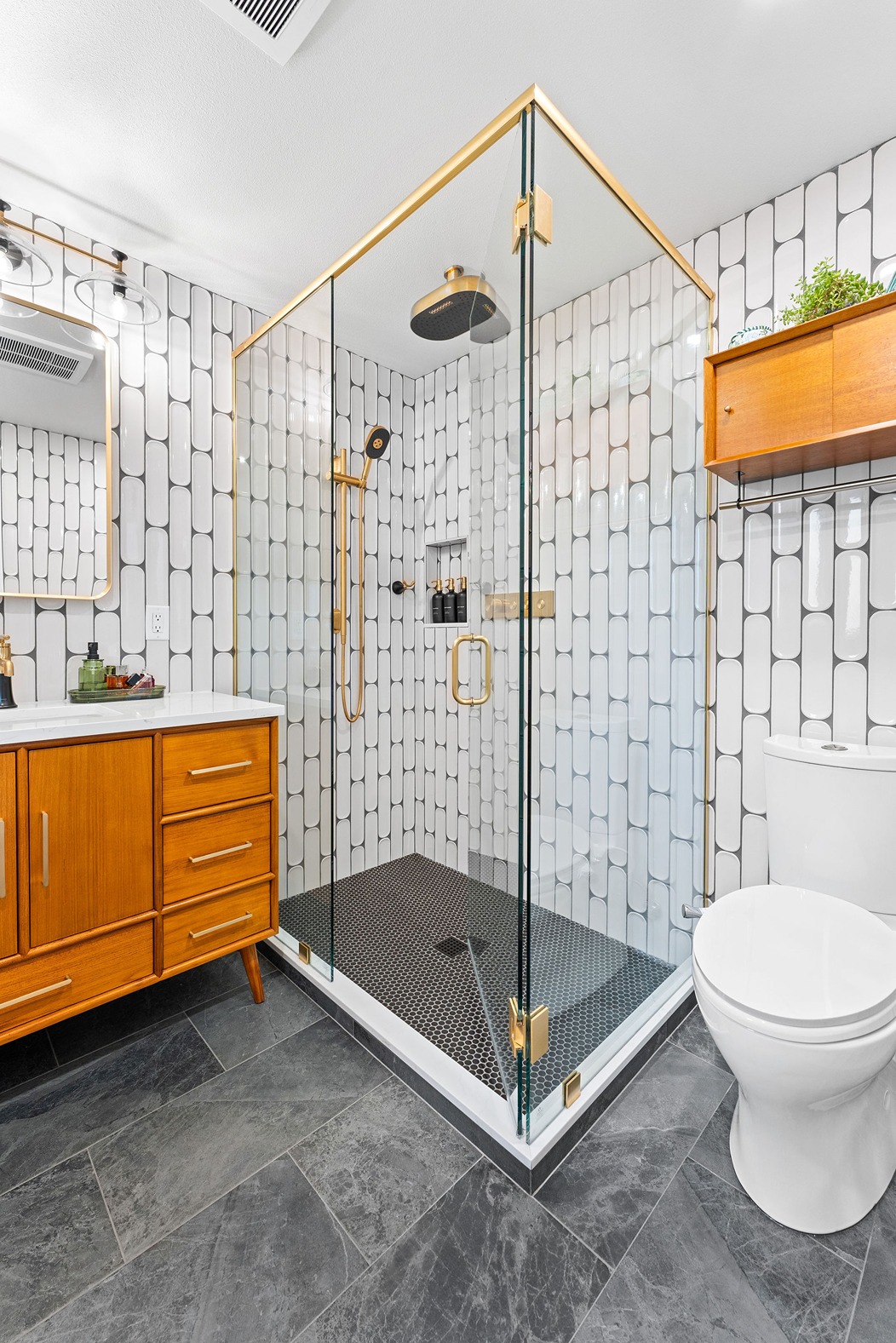 Modern bathroom with glass walk-in shower, gold fixtures, wood vanity, white tile walls, and dark stone flooring