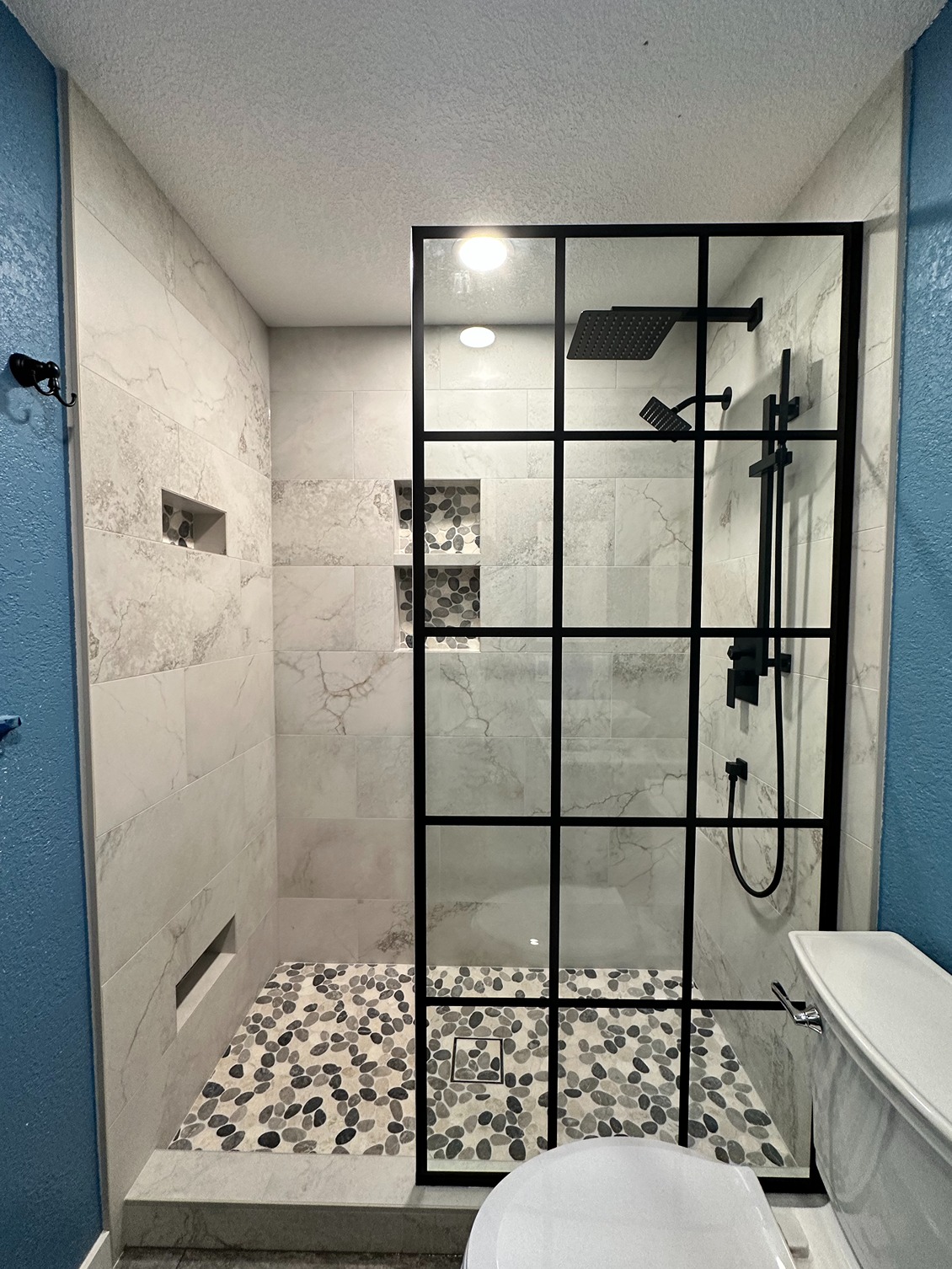 Modern bathroom with walk-in shower featuring black grid glass doors, matte black fixtures, marble-look tile walls, and pebble stone flooring