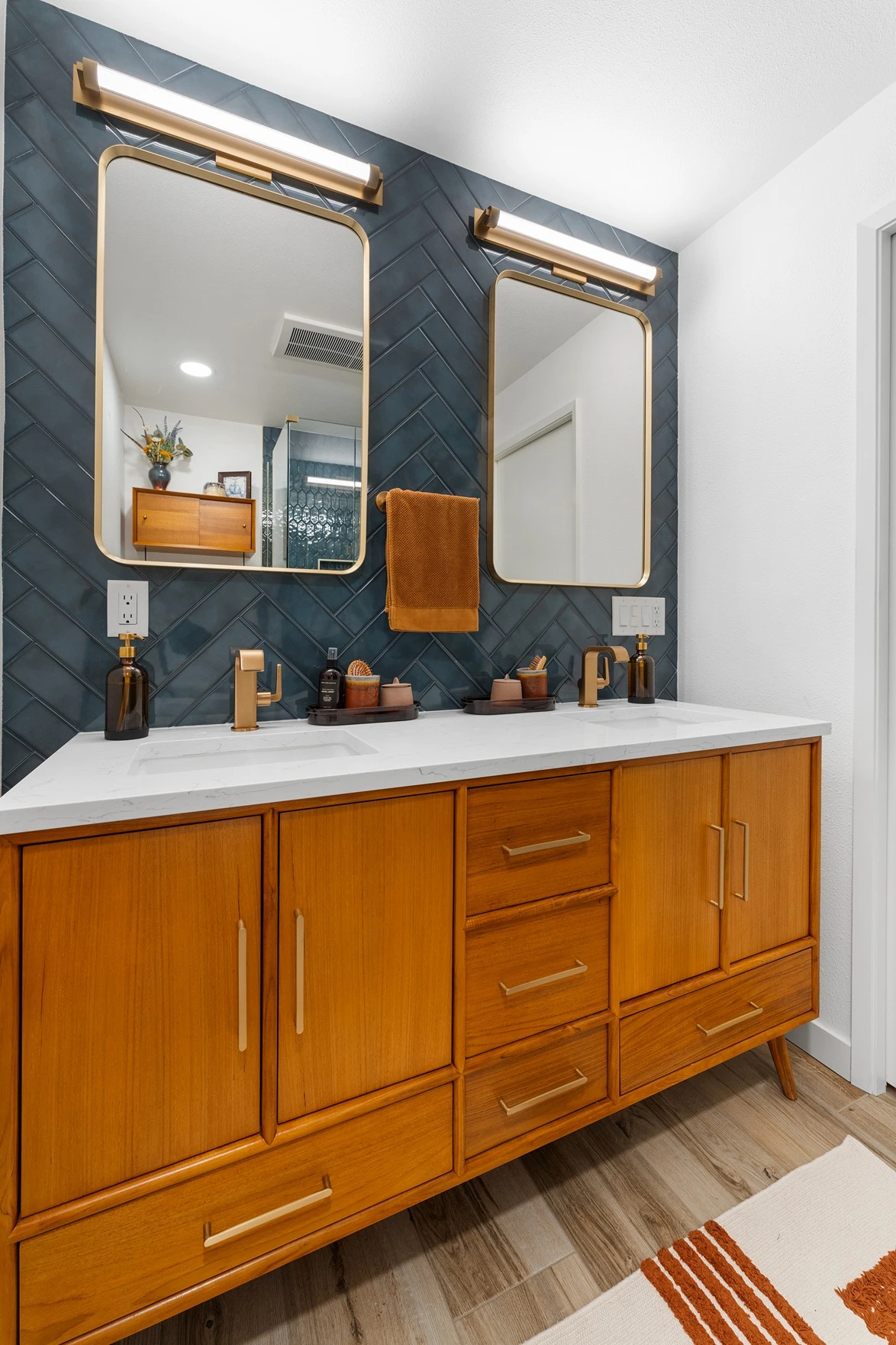 Teak double sink bathroom vanity with gold faucets, rounded brass-framed mirrors, LED bar lighting, and dark teal chevron wall tile.