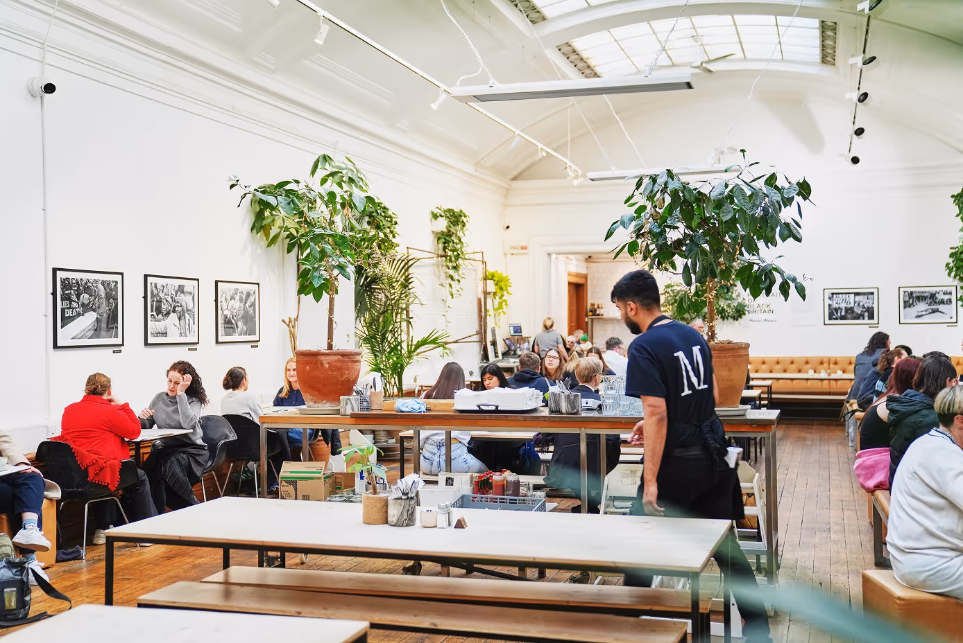 Medicine New Street. A bright, spacious café with wooden floors, people seated at tables, large potted plants, and a waiter in a black shirt.