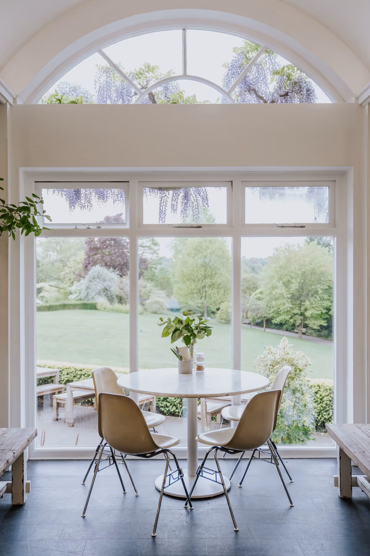 Medicine at Botanical Gardens in. Birmingham. A round white table with a small plant centerpiece surrounded by four chairs near large windows overlooking a green garden.