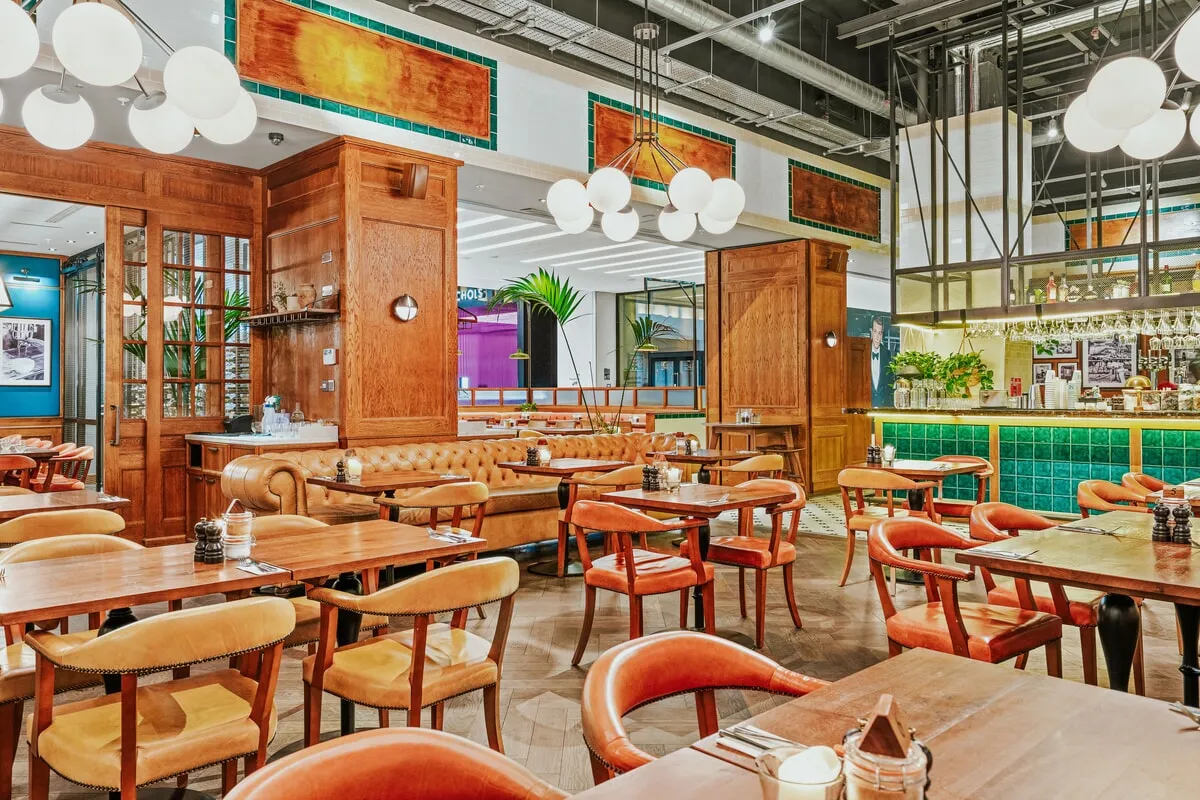 Bright restaurant interior with wooden tables, leather chairs, tufted booth seating, hanging globe lights, and a bar with green tiled front.