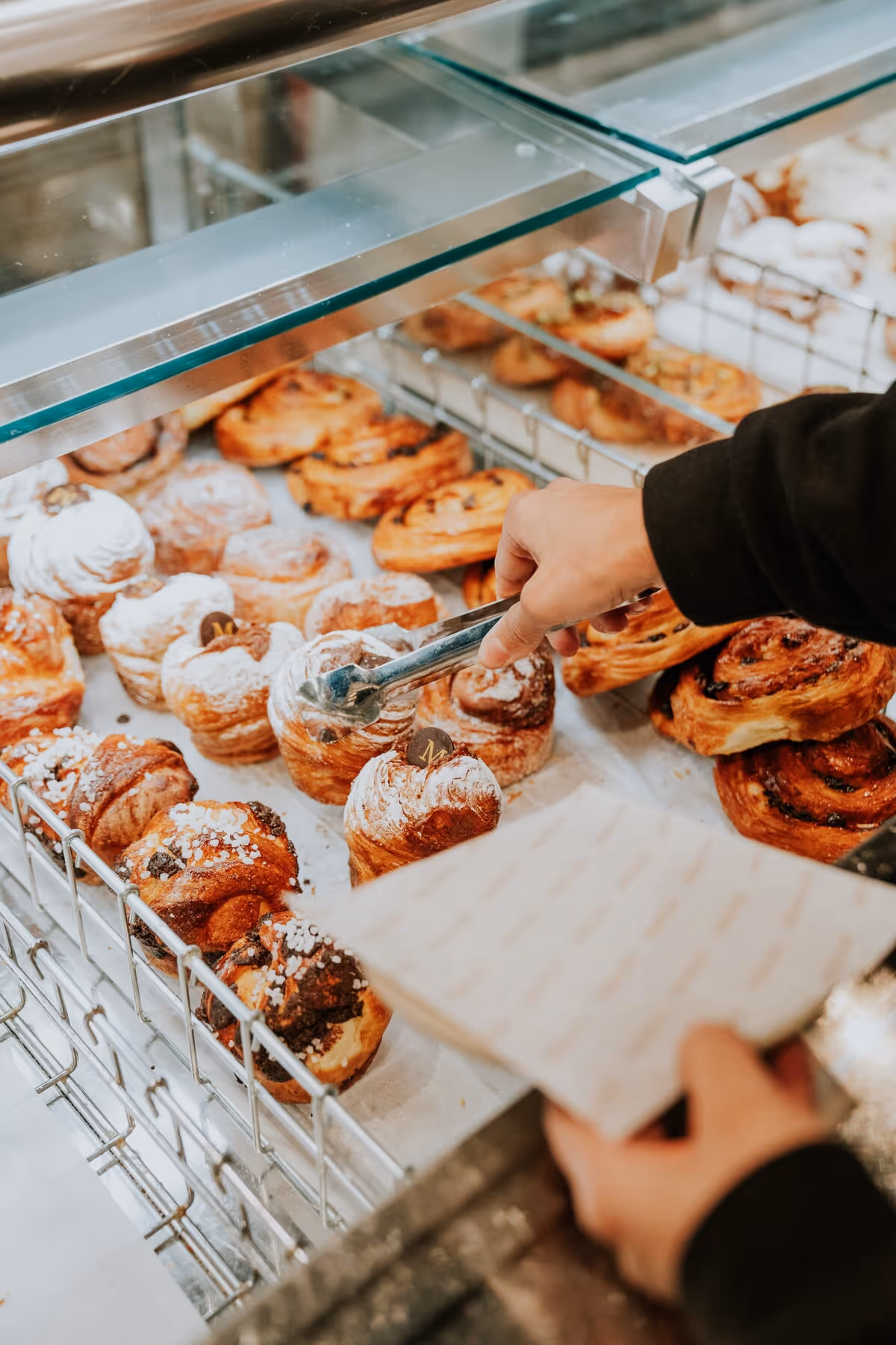Hand using tongs to pick a pastry from a bakery display case filled with various pastries.
