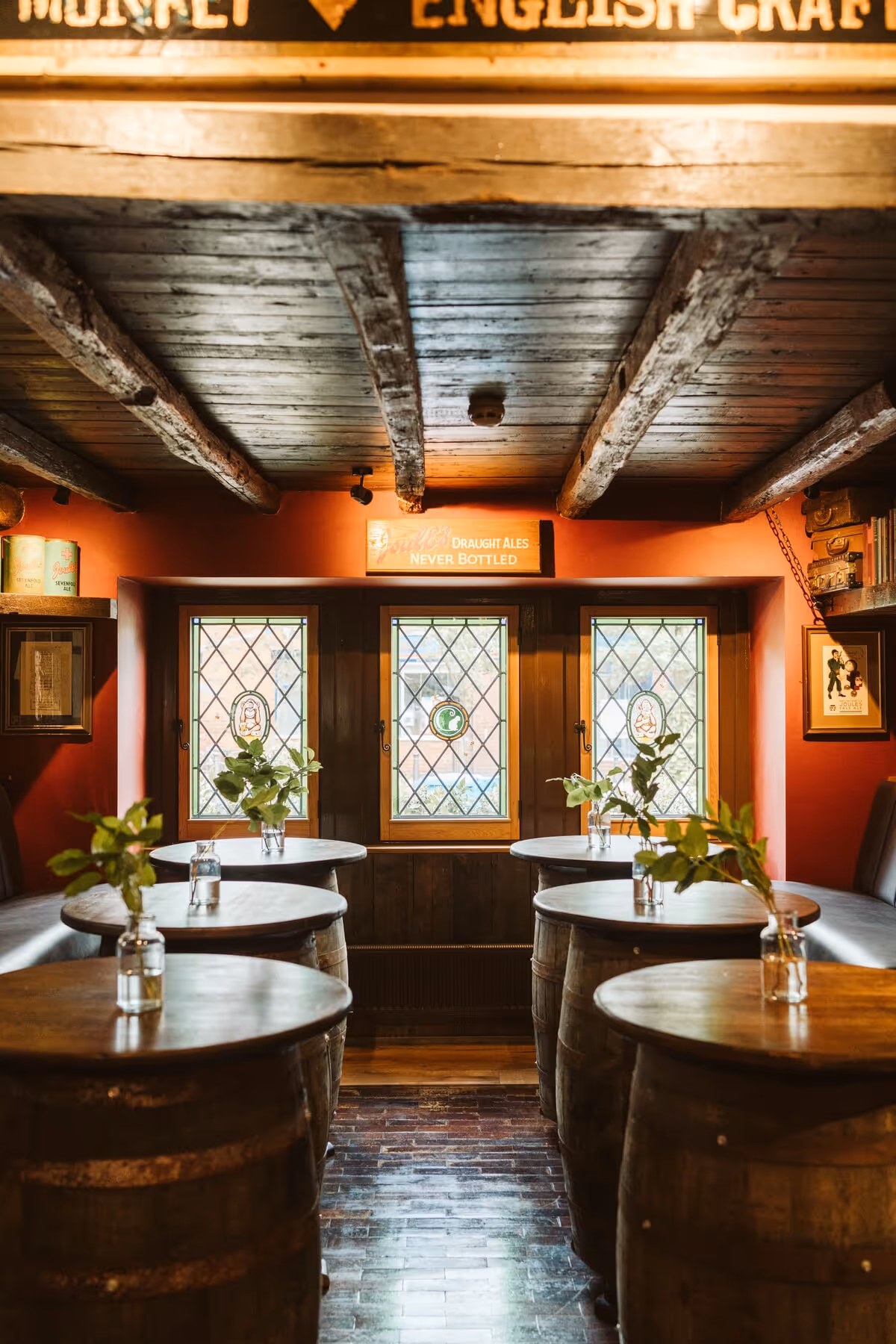 Cozy pub interior with wooden barrel tables topped with small vases of green plants, stained glass windows, and exposed wooden ceiling beams.