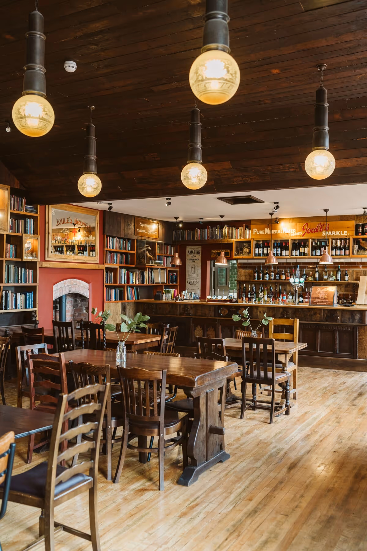 Cozy pub interior with wooden tables and chairs, bookshelf walls, pendant lights, and a bar stocked with bottles and taps.
