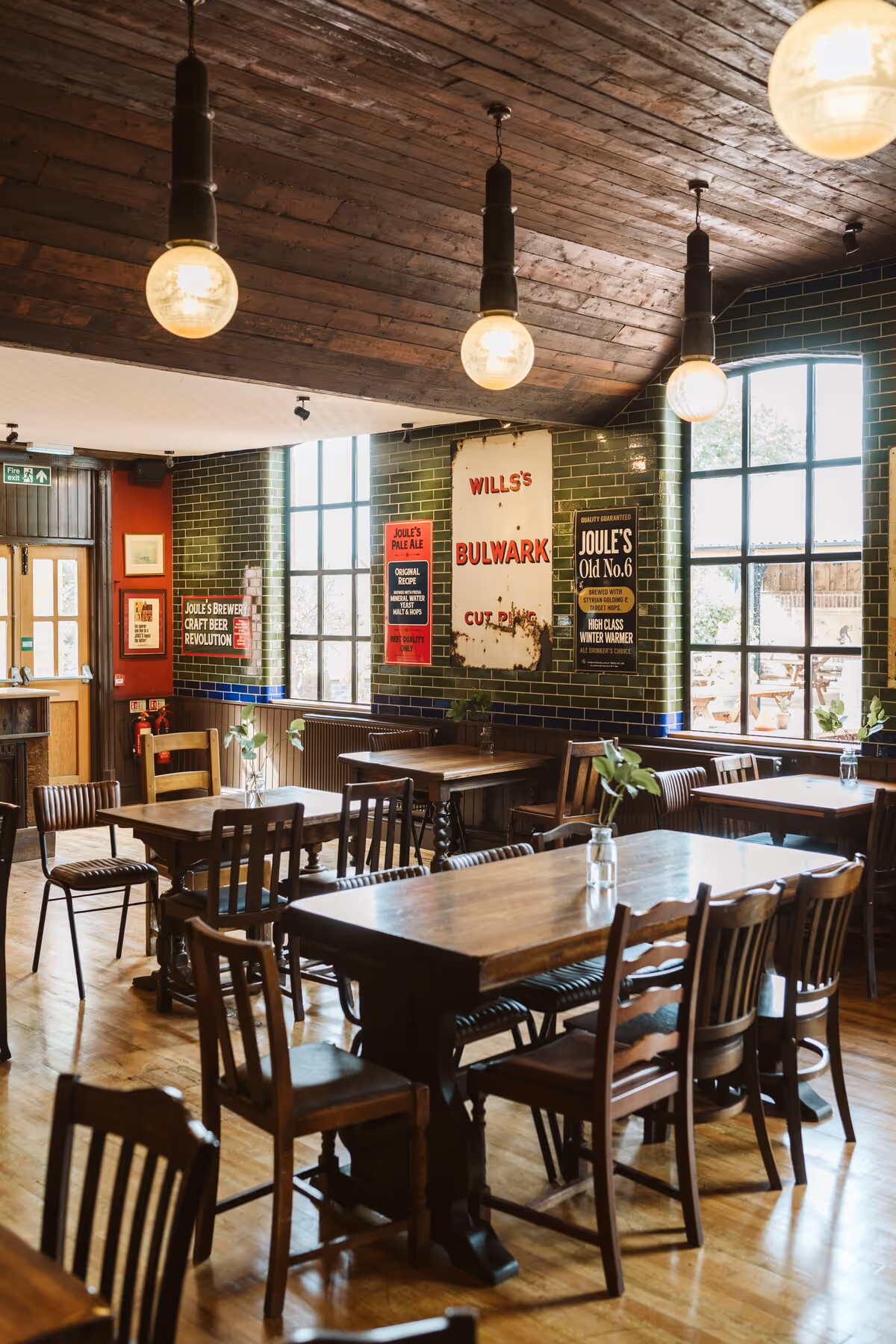 Cozy pub interior with wooden tables and chairs, hanging light bulbs, and large windows letting in natural light.