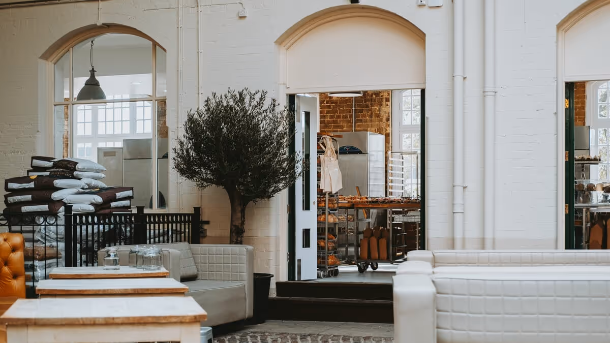 Modern bakery seating area with white and tan couches and a view into the bakery kitchen through an open doorway.