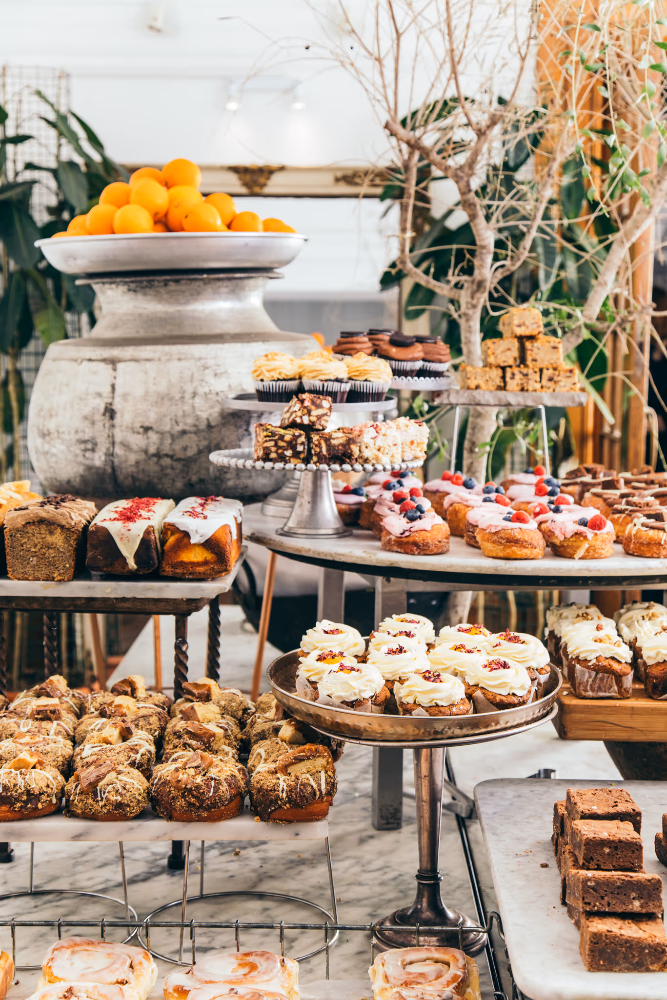 Display of assorted baked goods including cupcakes, brownies, donuts, and cakes arranged on metal stands and marble tables in a bakery setting.