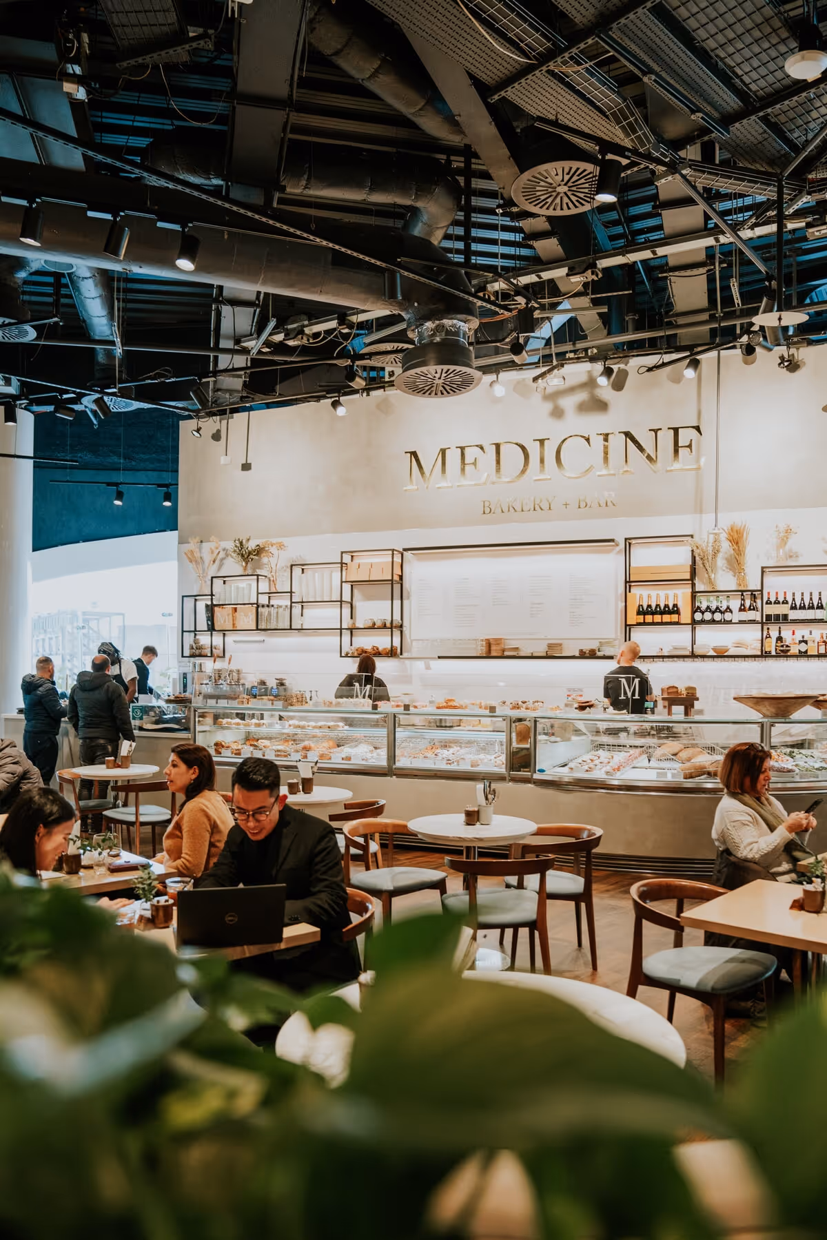 Interior of a modern bakery and bar named MEDICINE with customers seated at tables and staff behind the counter.