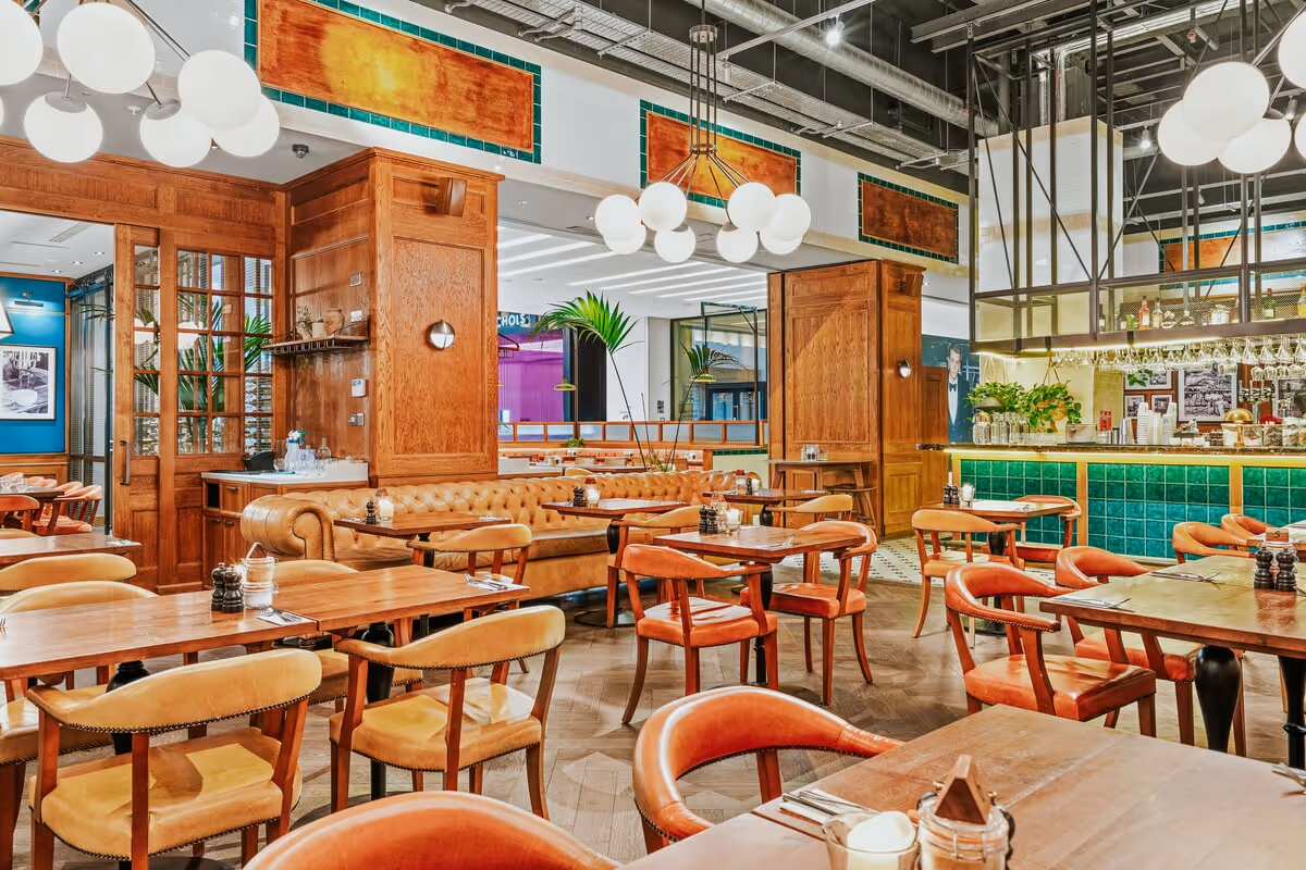 Bright restaurant interior with wooden tables, leather chairs, tufted leather banquette, and green-tiled bar.