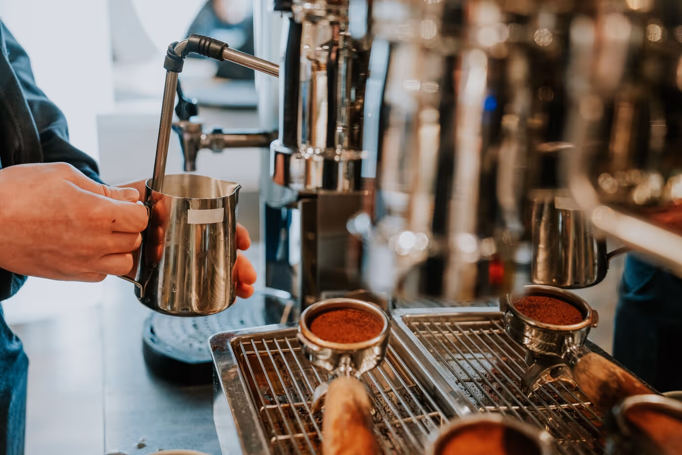 Person holding a metal milk frothing pitcher under a steam wand of an espresso machine with coffee grounds in portafilters nearby.