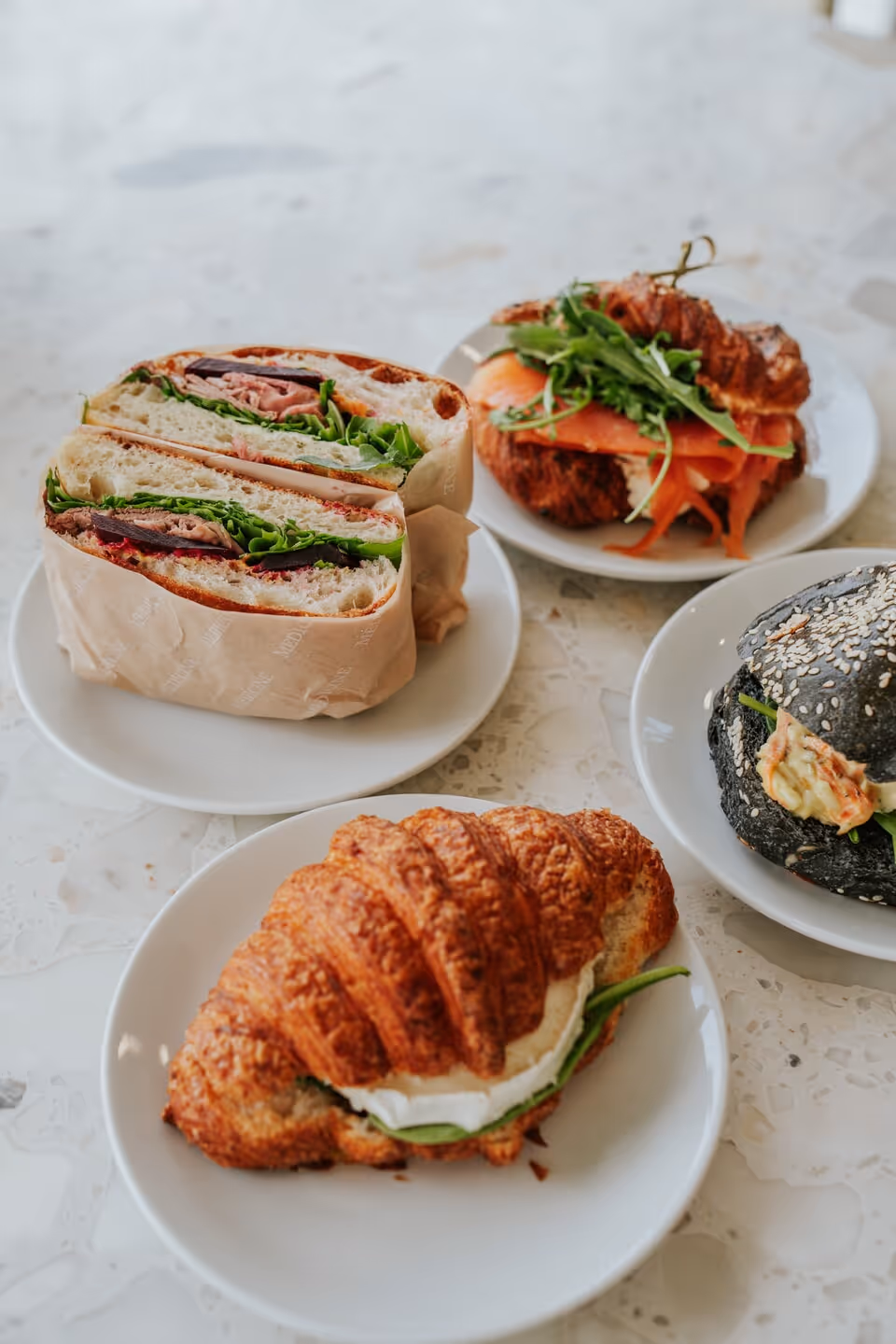 Assorted sandwiches including a croissant with cream cheese and greens, a sandwich with lettuce and meat, and a black sesame seed bun sandwich on white plates.
