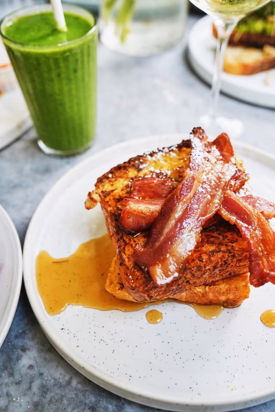 Plate with two slices of French toast topped with crispy bacon and drizzled with syrup, next to a green smoothie.
