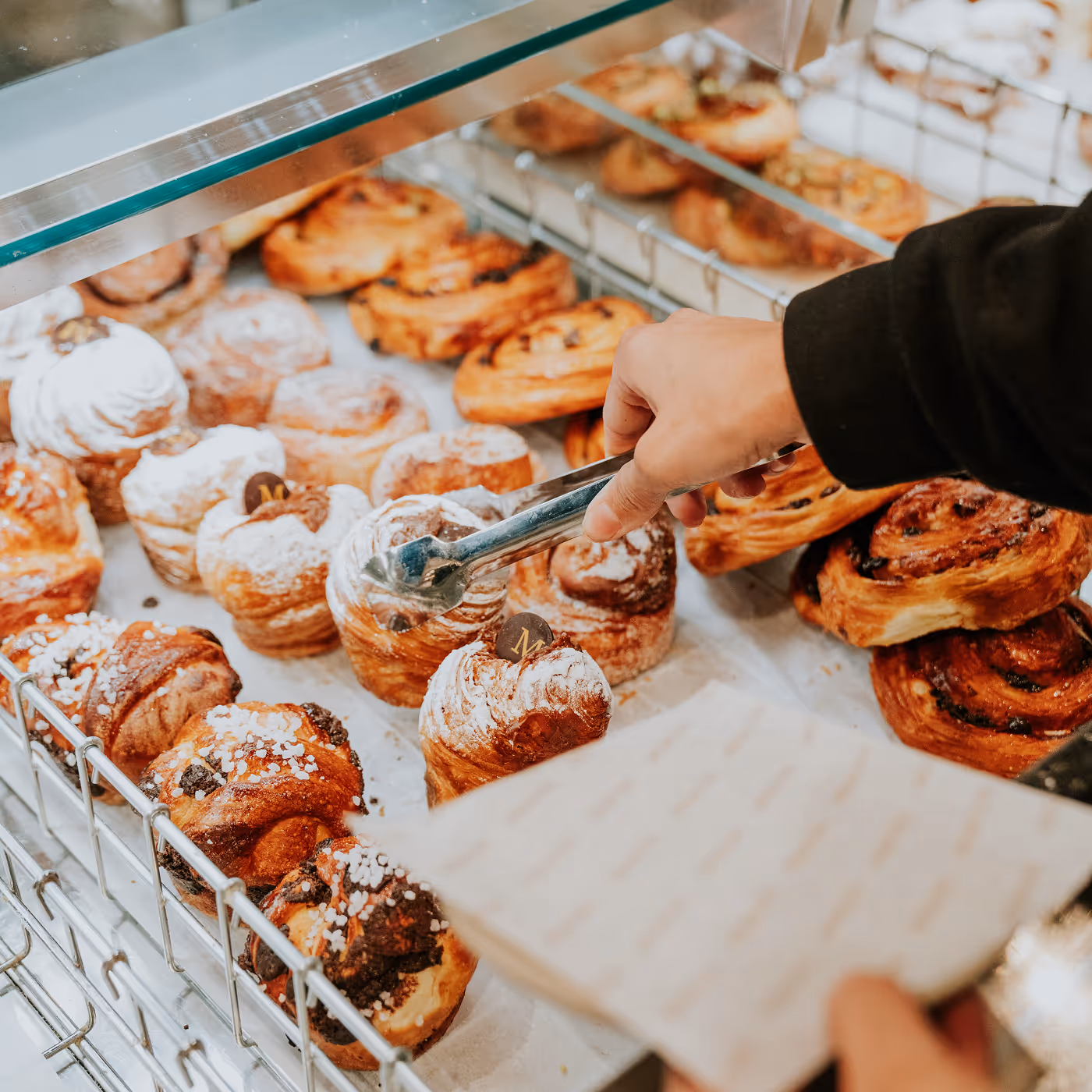 Hand using tongs to pick a powdered pastry from a display case filled with assorted baked goods.