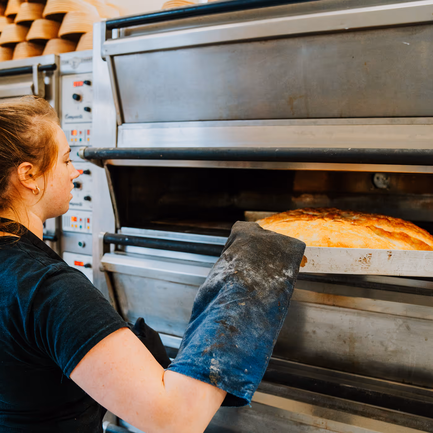 Baker wearing oven mitts pulling a large loaf of bread out of an industrial oven.