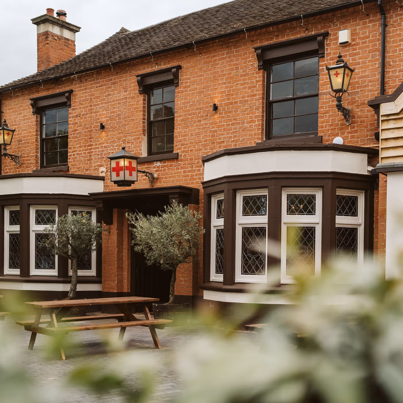 Brick building with brown framed windows and red cross lanterns above the entrance, with a wooden picnic table and small olive trees outside.