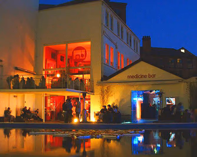Night view of a lively bar with brightly lit red and blue interiors, people socializing inside and outside by a reflective pool.