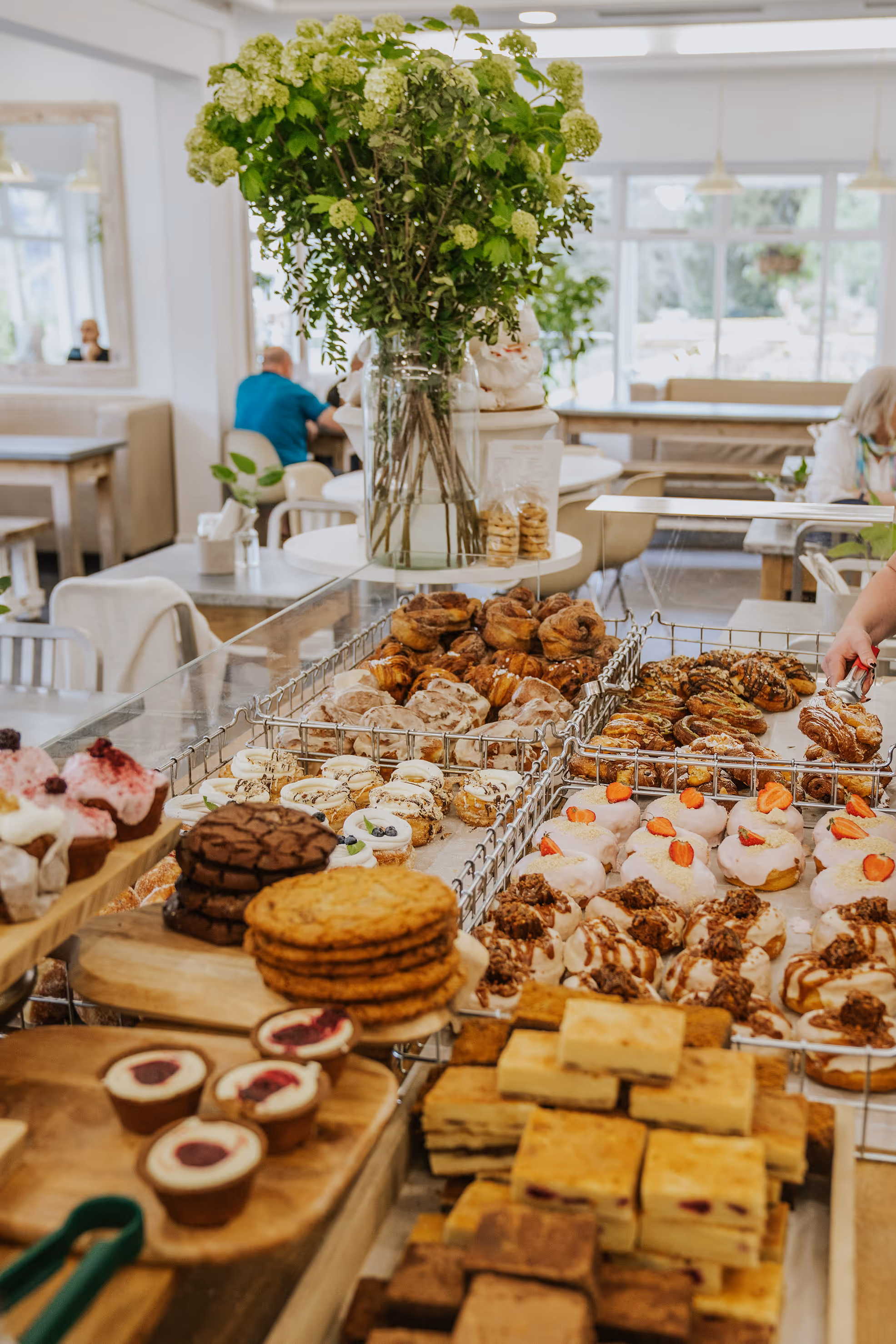 Display of assorted pastries, cookies, and desserts in a bakery with a large vase of green flowers in the background.