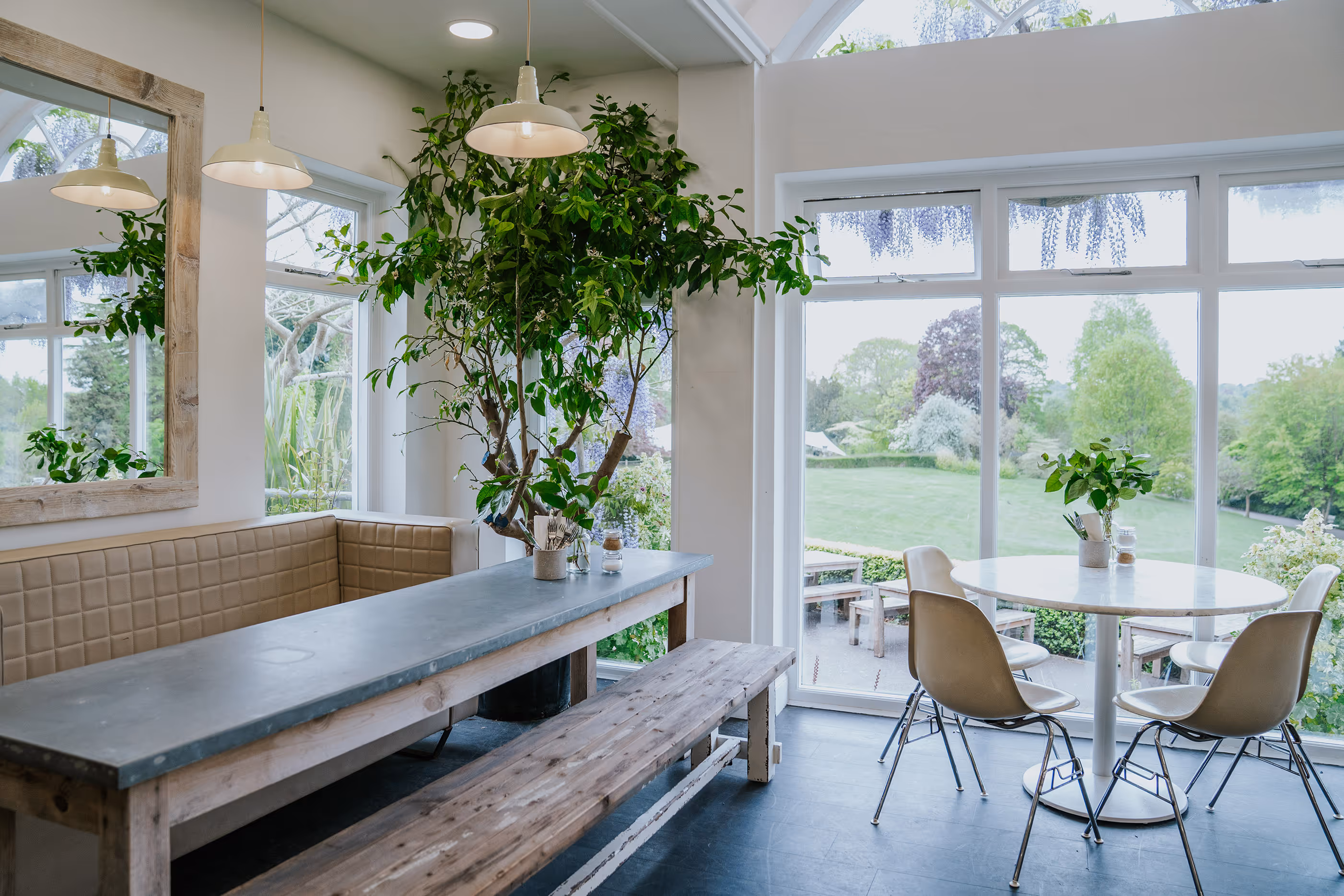 Bright dining area with a long gray table, wooden bench, beige cushioned seating, and a round table with chairs by large windows overlooking a green garden.