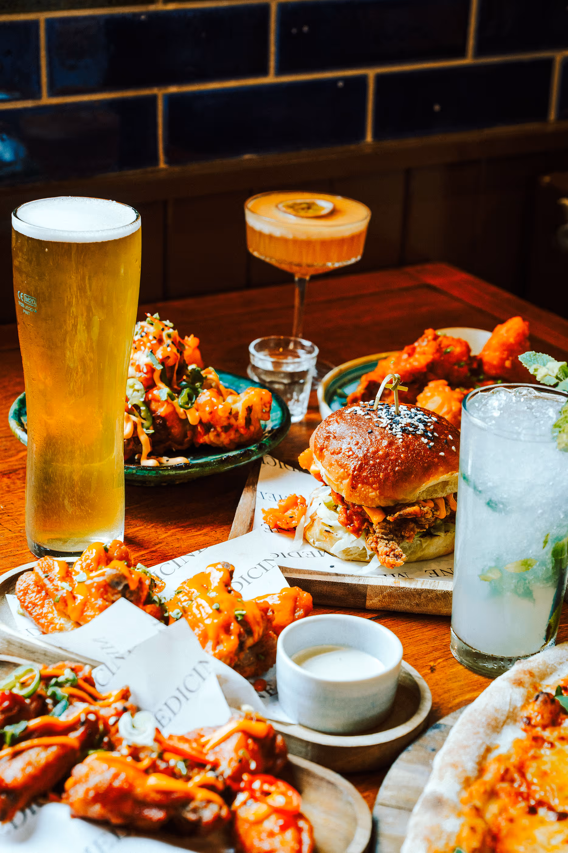 Table with a pint of beer, cocktail, iced drink, fried chicken sandwich, buffalo wings with dipping sauce, and a pizza slice.