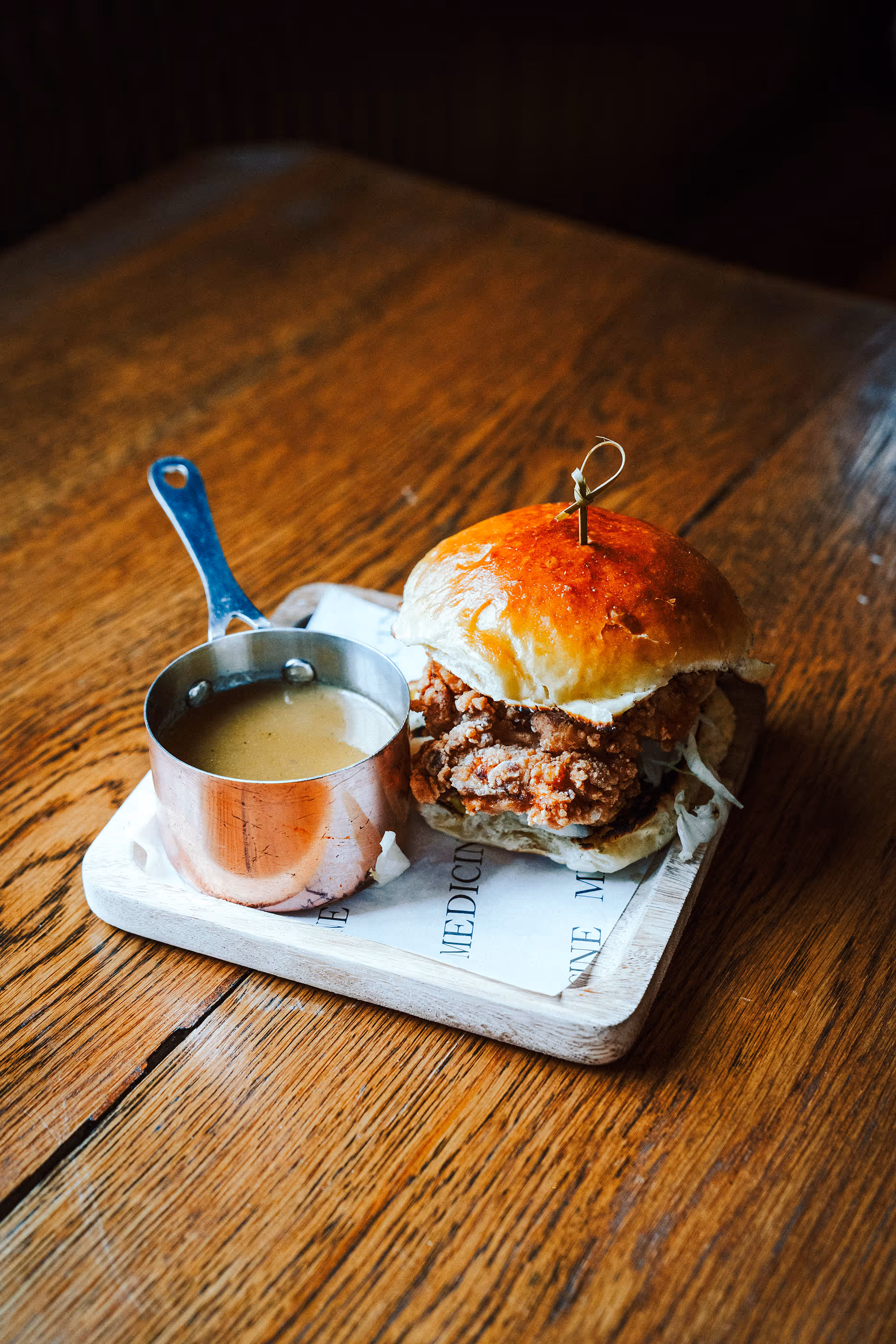 Fried chicken sandwich with lettuce in a glossy bun, served with a small metal cup of sauce on a wooden board.