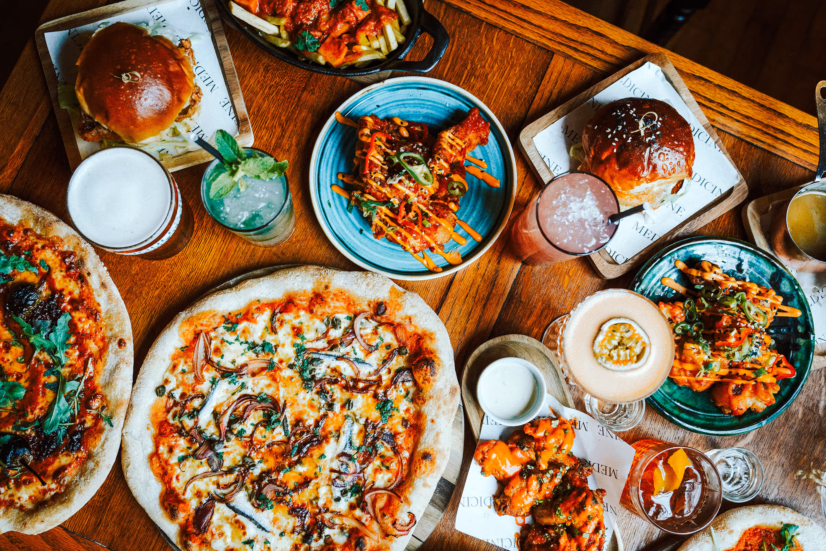 Overhead view of a wooden table with pizzas, burgers, sauced chicken wings, fries, and an assortment of cocktails and beers.