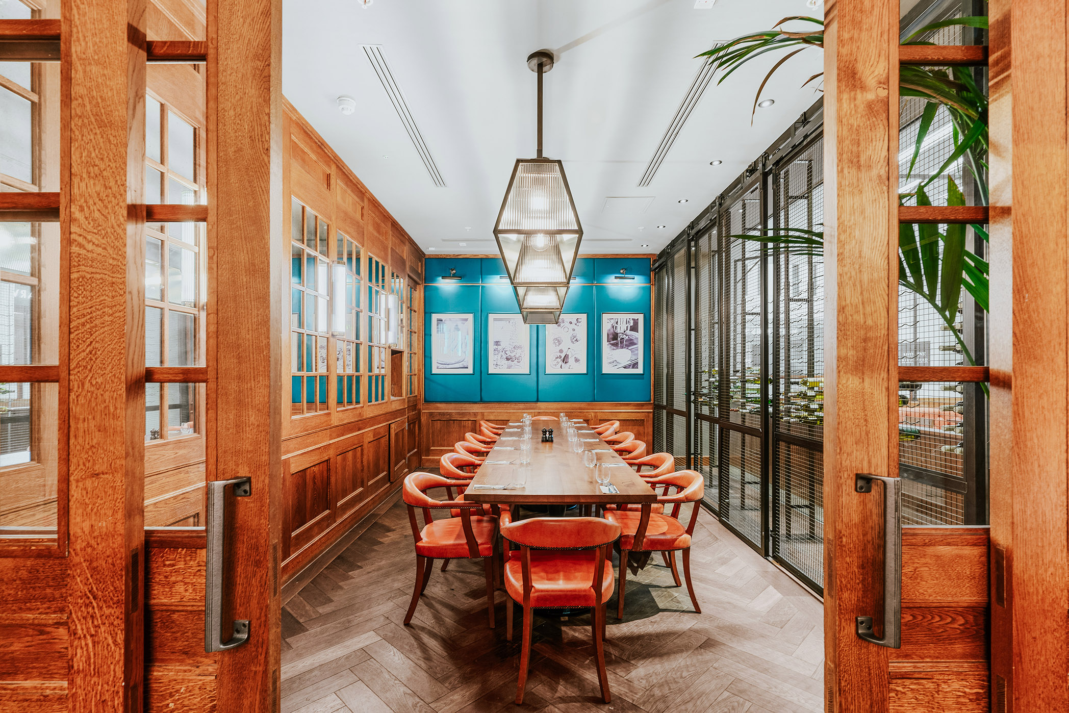 Long wooden dining table with orange leather chairs in a room featuring wood paneling and teal accent wall with framed artwork.