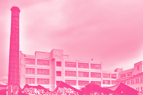 Historic industrial building with a tall brick chimney and graffiti-covered structures in the foreground under a cloudy sky.