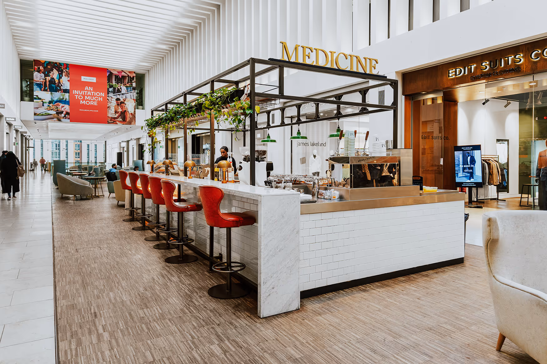 Modern indoor cafe with a marble counter, red bar stools, hanging plants, and a sign reading 'MEDICINE' above the counter in a shopping mall corridor.