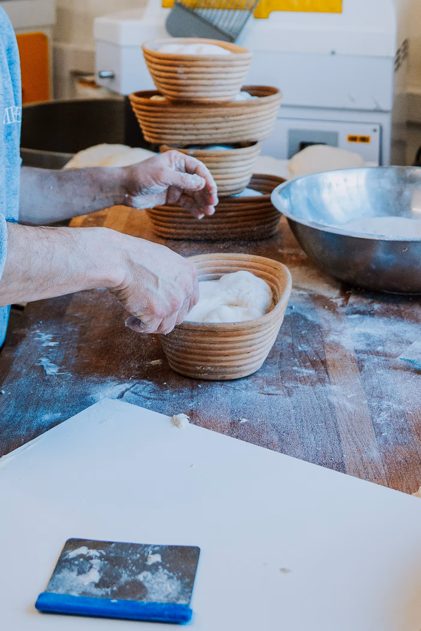 Person shaping dough in a round wicker basket on a flour-covered wooden table with baking tools and stacked baskets in the background.