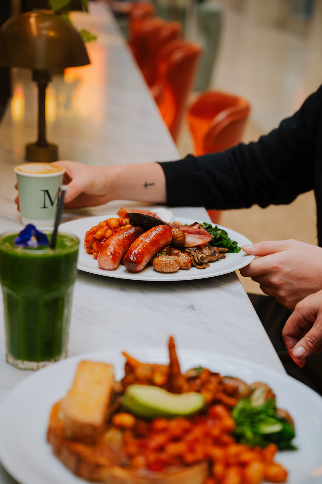 Person holding a plate with sausages, baked beans, mushrooms, and greens at a marble counter with a green smoothie and coffee cup.