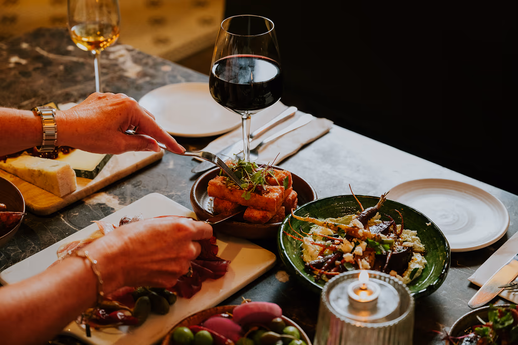 Person using fork and knife to eat plated gourmet food with a glass of red wine on a marble table.