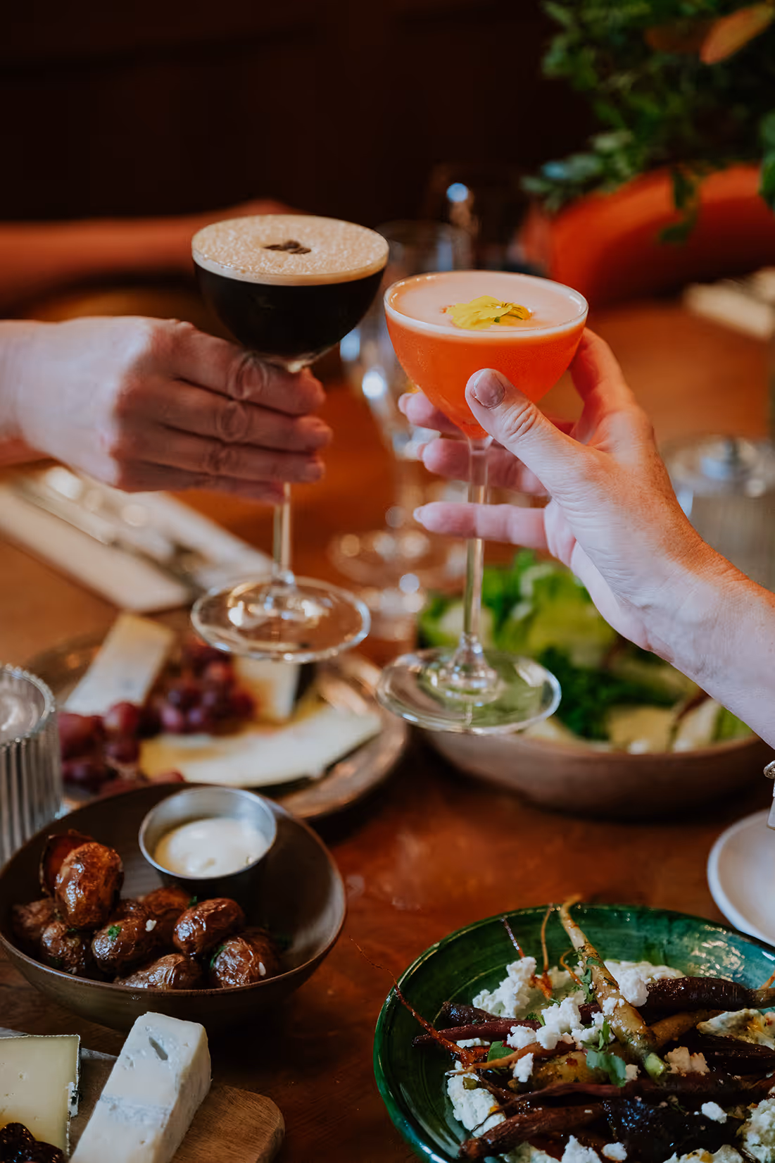 Two hands clinking cocktail glasses over a table with roasted potatoes, cheese, salad, and a vegetable dish.