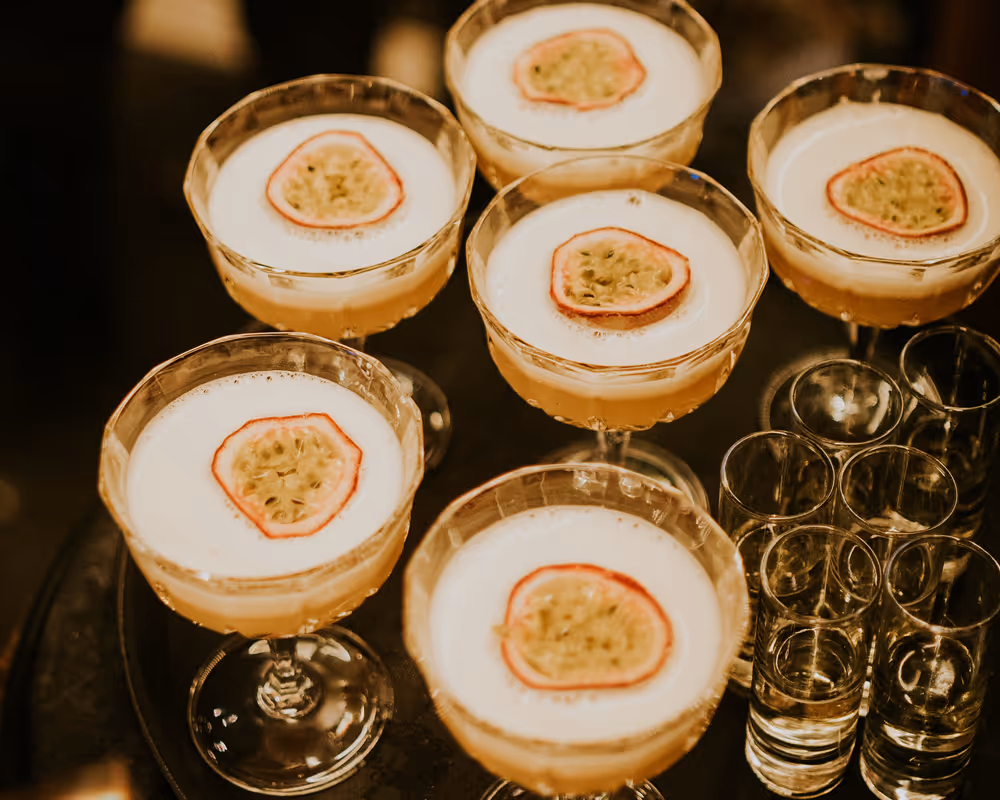 Six coupe glasses filled with frothy cocktails garnished with passion fruit slices on a tray.