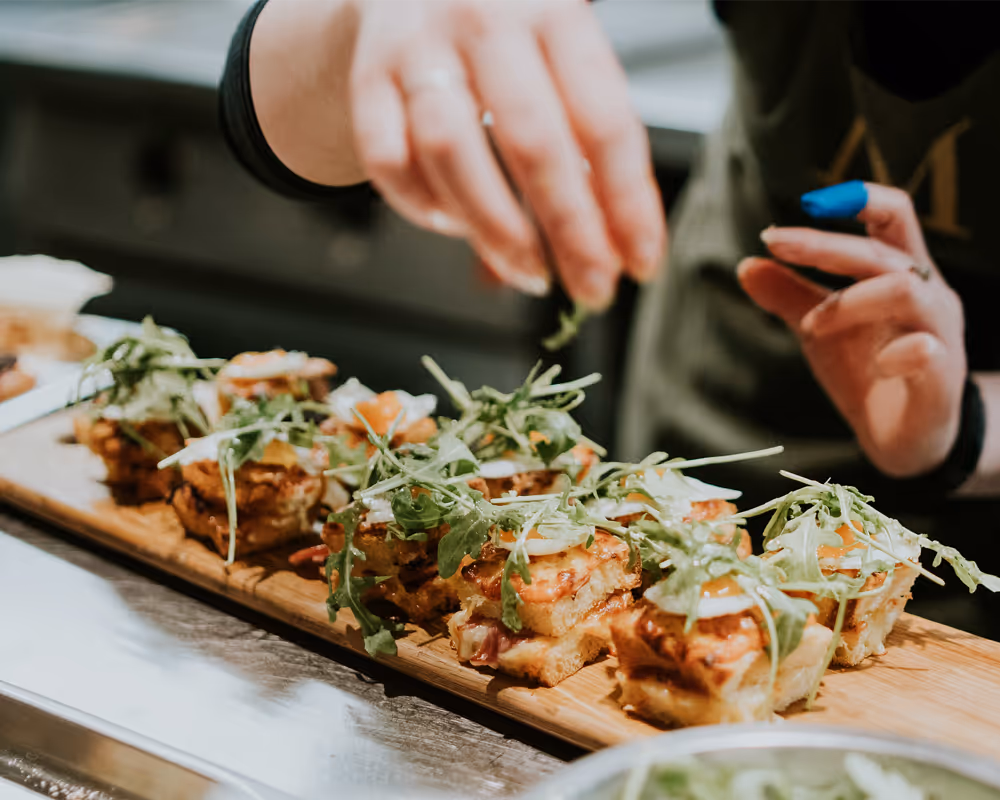Close-up of a person garnishing bite-sized appetizers with greens on a wooden serving board.