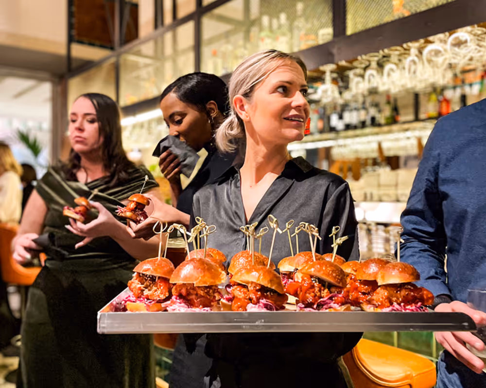 Server holding a tray of small fried chicken sliders with coleslaw in a busy restaurant.