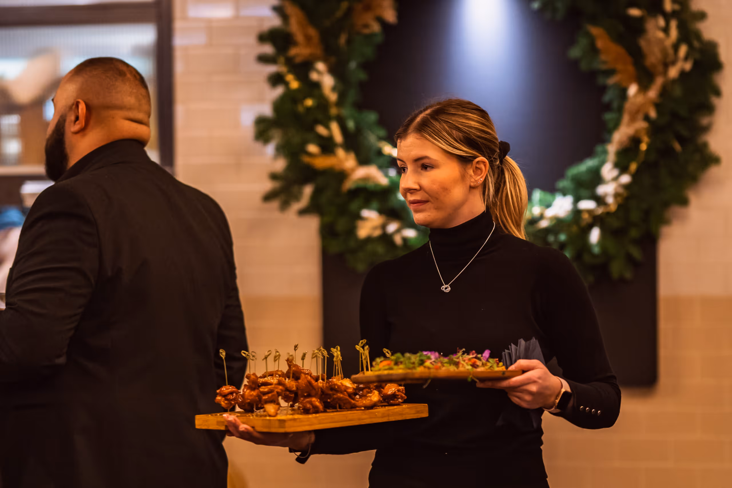 Woman in black turtleneck carrying wooden trays with skewered appetizers and finger foods at an event.