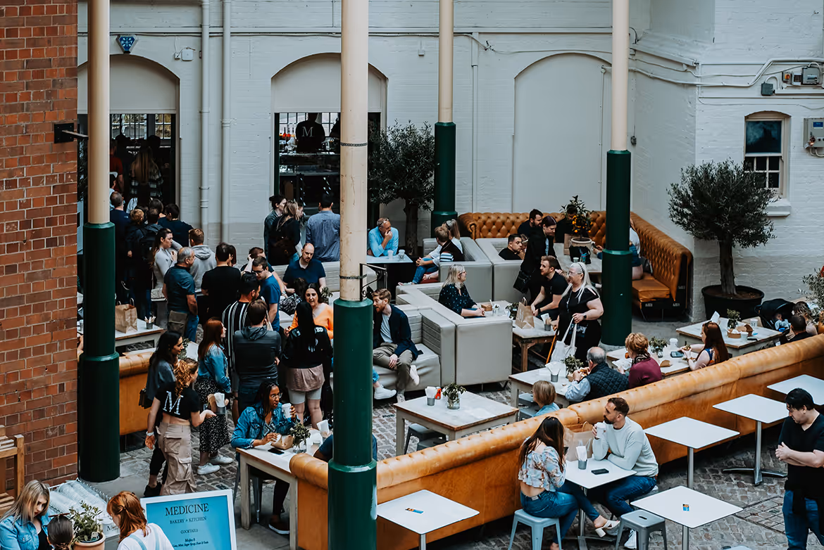 Crowded indoor cafe with people sitting at tables and standing in line, surrounded by white walls and green pillars.