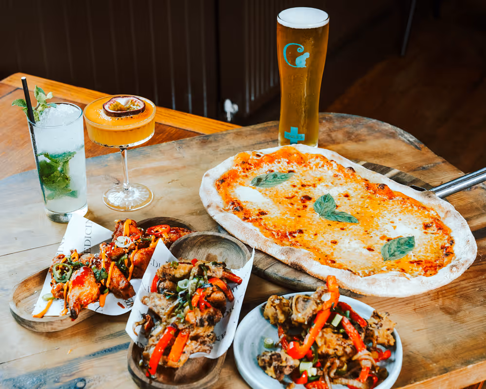 Wood-fired pizza with basil leaves, spicy chicken wings, a tall glass of beer, a cocktail with passion fruit, and a mojito on a wooden table.