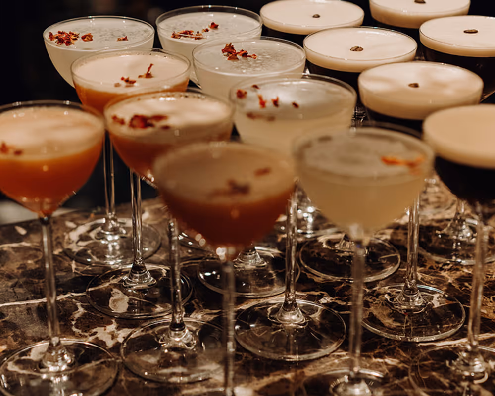 Multiple cocktail glasses filled with frothy drinks garnished with dried flower petals and coffee beans on a marble surface.