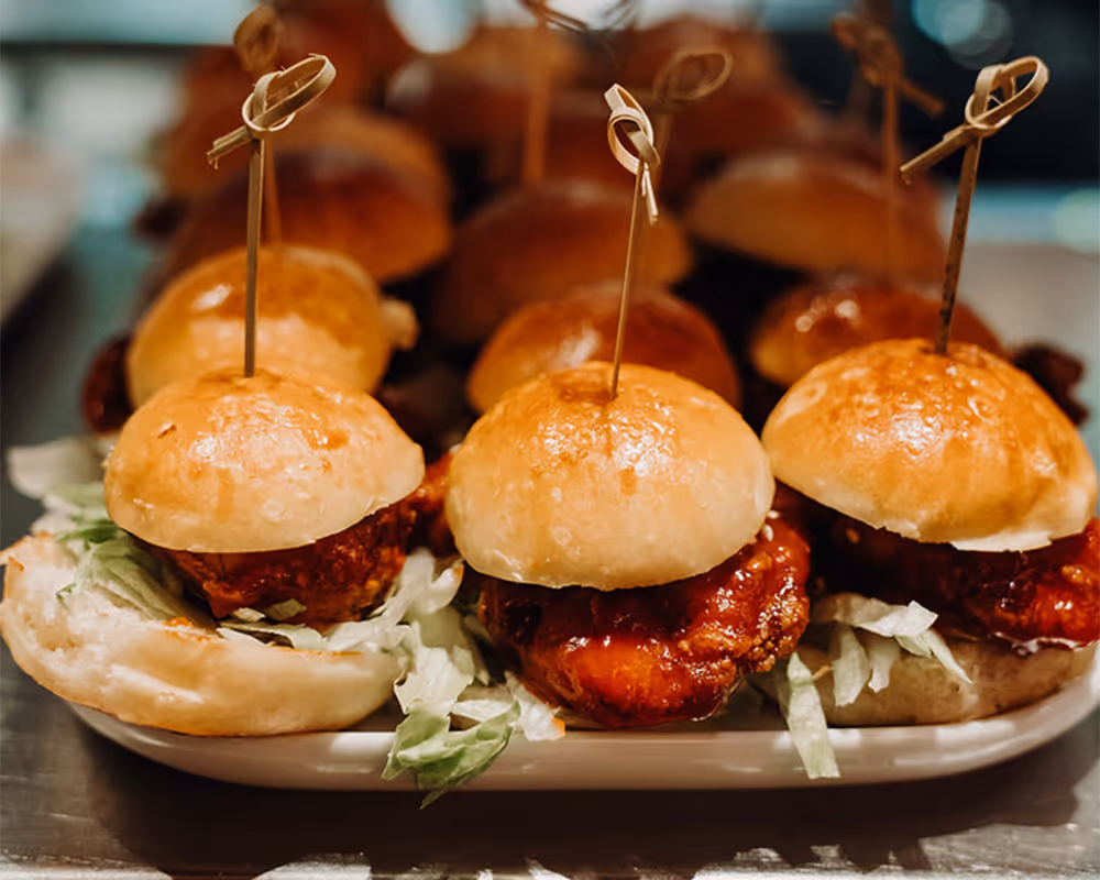 Tray of fried chicken sliders with lettuce on small buns, each secured with a bamboo skewer.