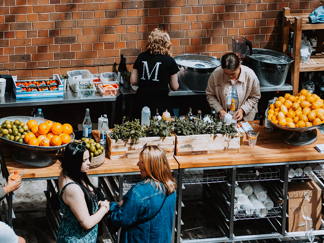 People preparing drinks behind a wooden counter with bowls of oranges and limes and containers of ingredients in a bright indoor setting.