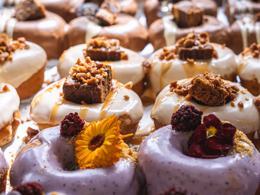 Close-up of assorted gourmet donuts topped with icing, brownie pieces, nuts, blackberries, and edible flowers.