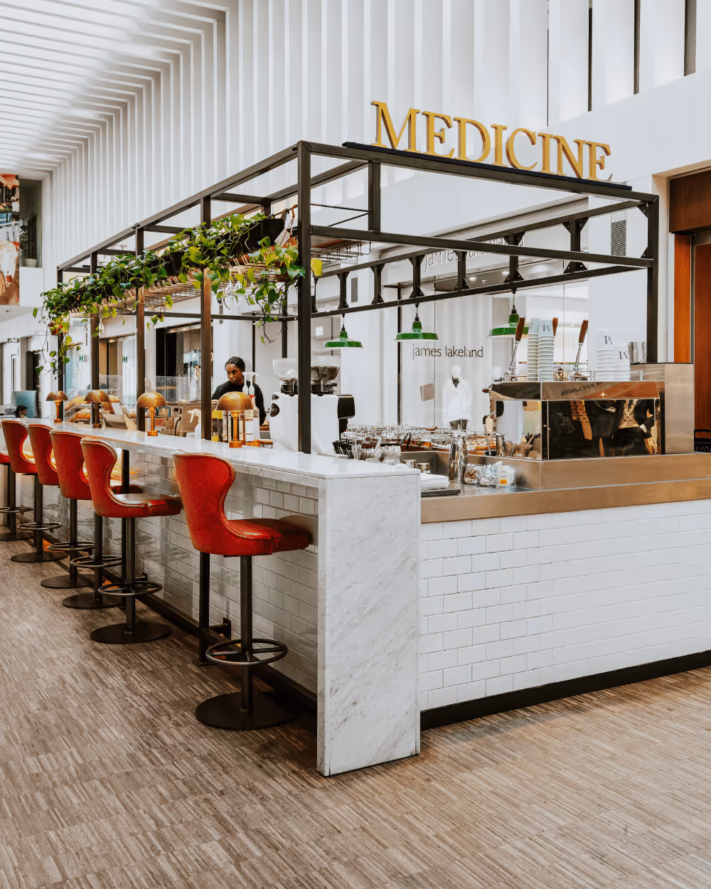 Modern cafe counter with red bar stools, green hanging plants, and a large 'MEDICINE' sign above.
